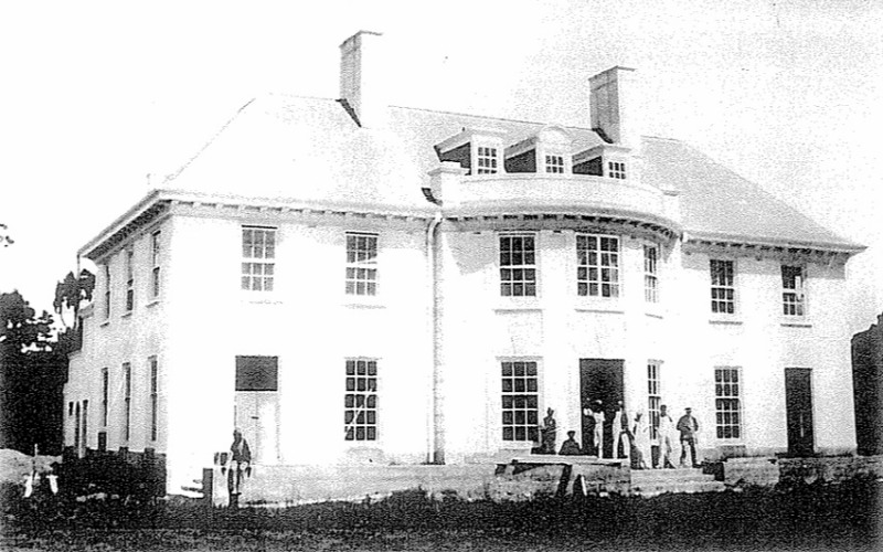 A black and white photo of a large white house with workers at the front.