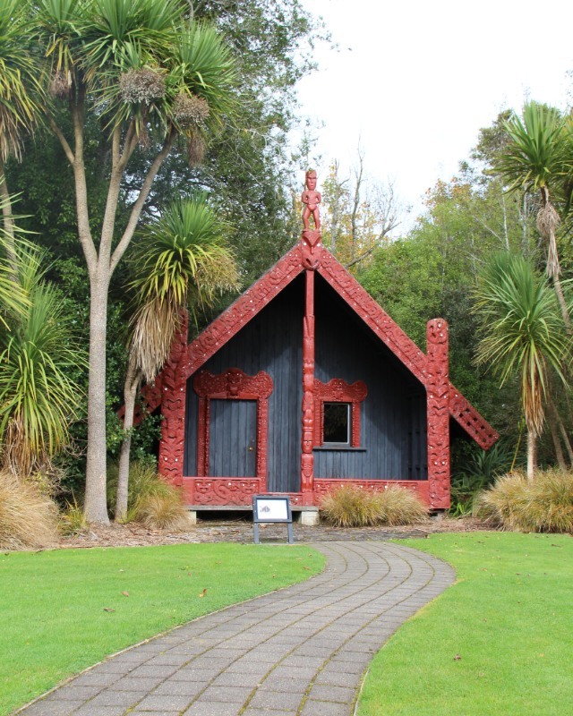 A carved Maori whare whakairo surrounded by trees with lawn in front.