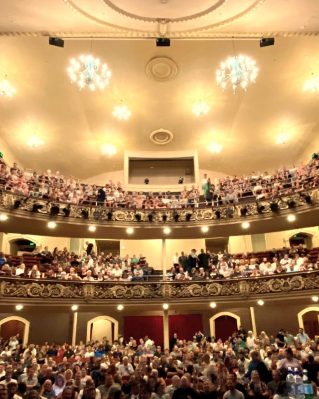 A crowd of people in a grand auditorium with chandeliers and gilded decorations.