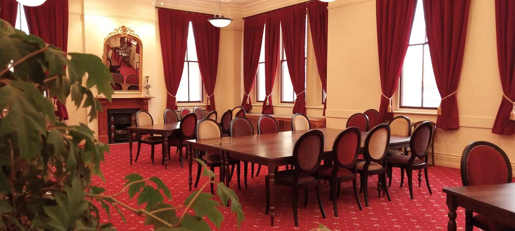 A high-ceiling room with a fireplace, wooden tables and chairs, red carpet and heavy red curtains.