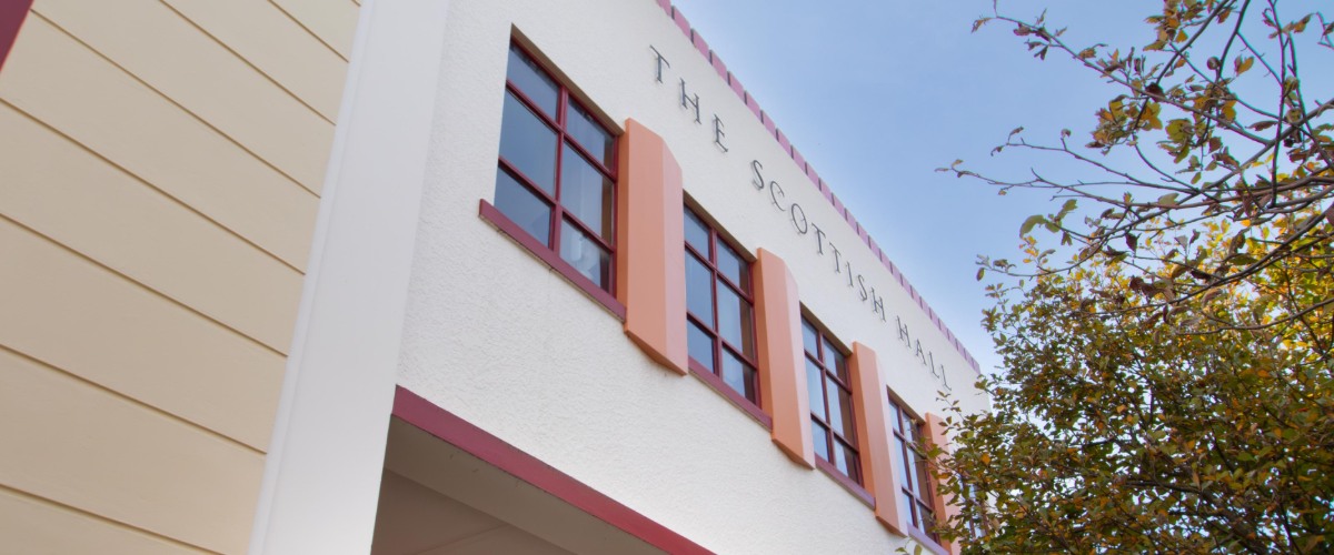 A cream and maroon building with letters on the front reading The Scottish Hall, a few trees in front.