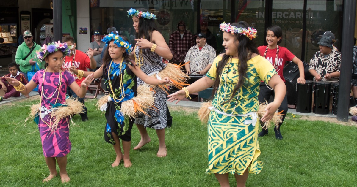 A group of four girls in traditional Polynesian dresses performing a dance on grass. They wear floral headbands and hold grass skirts, accompanied by musicians in the background. The setting appears to be an outdoor event.