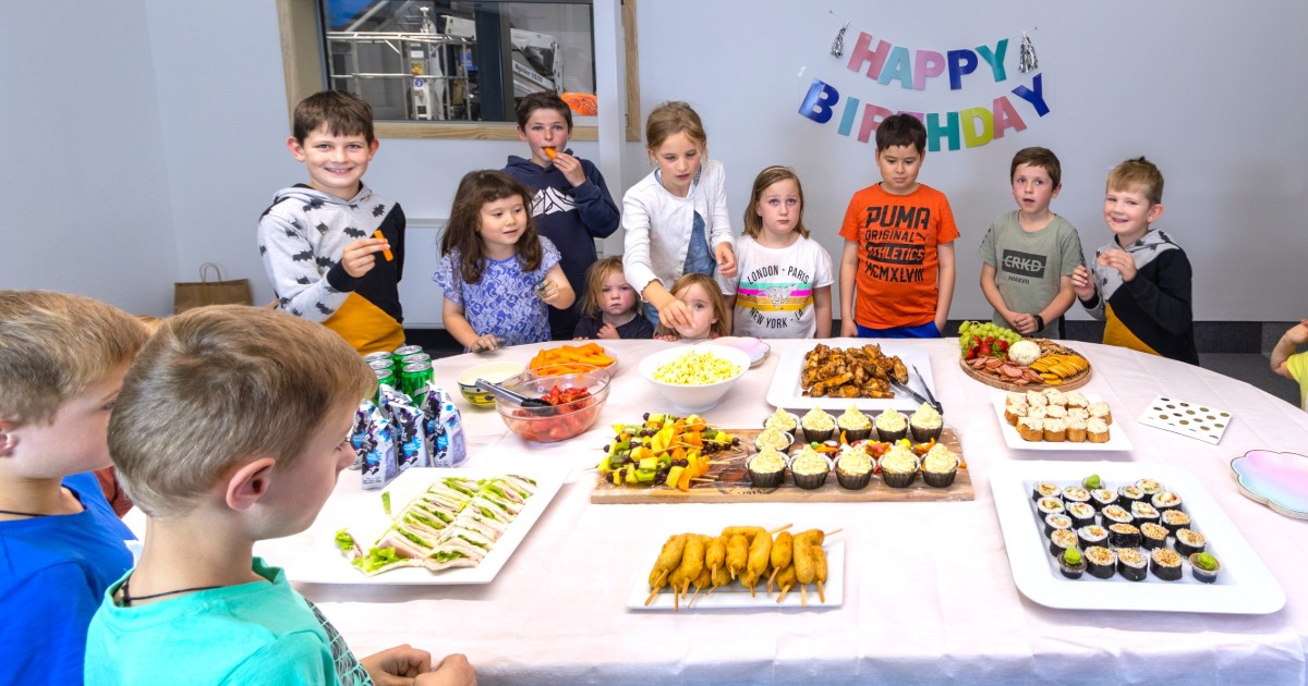 a  group of children around a table of party food with happy birthday banner on the wall.