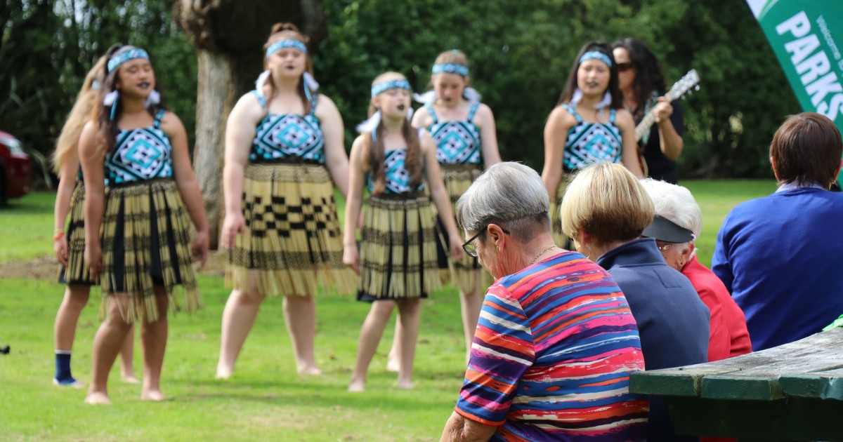 A group of young women in traditional attire, consisting of patterned tops and grass skirts, perform on a grassy field. A small audience sits on a park bench, watching the performance. A musician with a guitar stands among the performers.