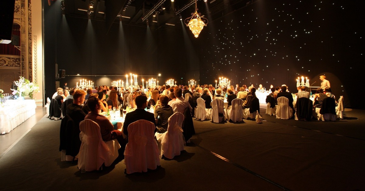 A large indoor event with elegantly dressed guests seated at tables, surrounded by soft lighting and candelabras. A decorated stage is in the background, adorned with a starry backdrop, creating a sophisticated atmosphere.