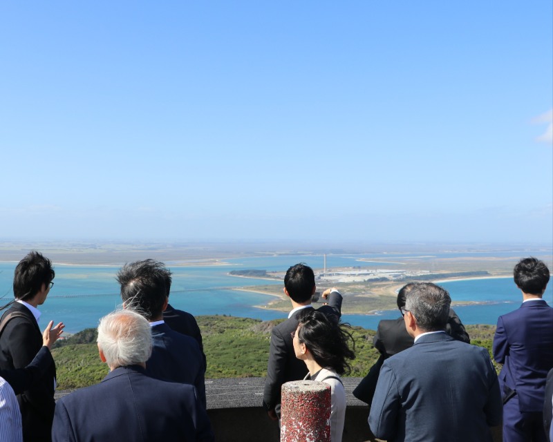 A group of people in formal attire, standing on a lookout point, observing a view of water and land. The scene includes a bright blue sky and a shoreline with industrial structures in the background. Some individuals appear to be taking photos.