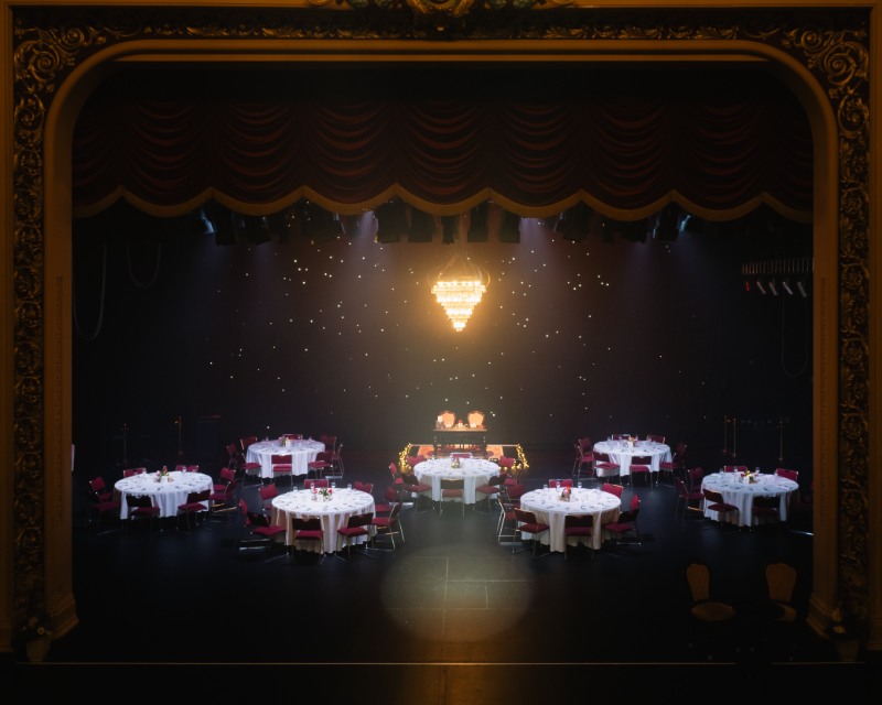 An elegantly set stage for a formal event featuring several round tables with white tablecloths and red chairs. A large chandelier hangs above, surrounded by a dark background with subtle lighting, creating a sophisticated atmosphere.