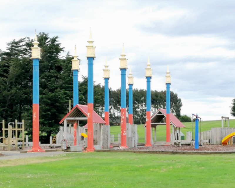 An outdoor playground featuring colorful tall poles with decorative tops, surrounded by grassy areas. The playground includes wooden structures and play equipment. Trees are visible in the background under a cloudy sky.
