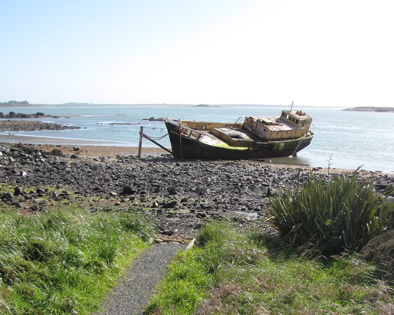 An abandoned fishing boat rests on a rocky shoreline, partially covered with green vegetation. The scene features calm water and distant islands under a clear sky, indicating low tide.